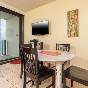 This cozy dining area features a round table, a TV, and a vibrant view of the beachfront from the balcony