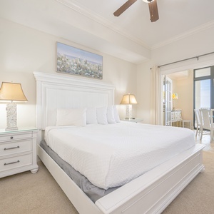 Master bedroom featuring a plush king bed and a balcony view with dining area and coastal artwork