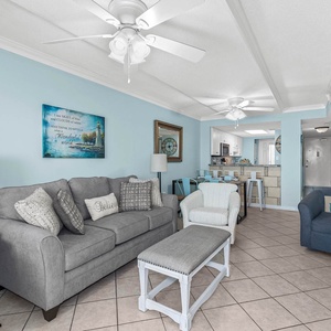 Living area featuring a cozy gray sofa with queen sleeper, beach-themed decor, and a view into the modern kitchen