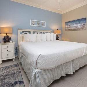 Brightly decorated bedroom features a king bed with plush pillows, a stylish nightstand, and coastal artwork on the walls