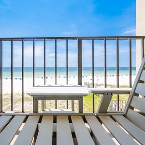 A comfortable balcony view features a table and chair overlooking a pristine beach with umbrellas dotting the white sand