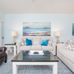Bright living area features a light-colored sofa with decorative pillows, a stylish coffee table, and vibrant coastal artwork