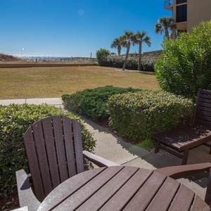 Relax on the patio with wooden seating, surrounded by lush greenery and a view of the beach in the distance
