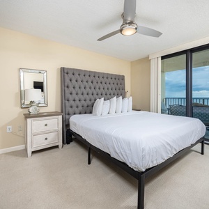 Cozy master bedroom featuring a plush king with gray tufted headboard, light bedding, and a balcony with a view of the beach