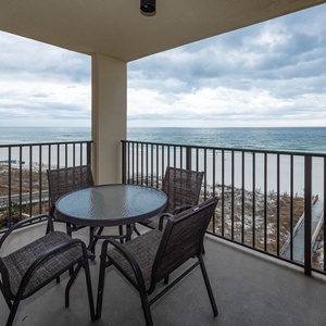 A cozy balcony features a round glass table and chairs, offering a serene view of the beach and gentle waves beyond