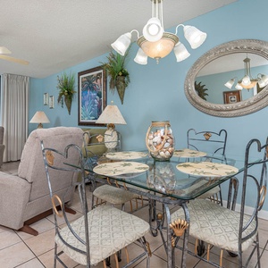 Dining area features a glass table with shell-themed placemats, surrounded by decorative chairs and a view of the gulf