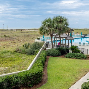 Lush green landscaping frames the inviting pool area, with lounge chairs and a view of the beach in the distance