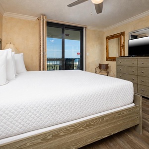 Elegant master bedroom featuring a king bed, dresser, TV, and access to a balcony with a view of the beach