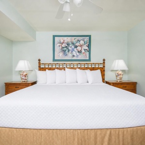 A bright bedroom features a large king bed with multiple pillows, complemented by wooden nightstands and a floral wall art