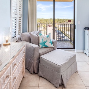 Cozy seating area features a light gray armchair with decorative pillows, a stylish dresser, and a view through balcony doors