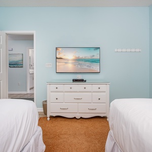 Bright guest bedroom featuring two double beds, a dresser with a TV, and cheerful beach artwork on soft blue walls