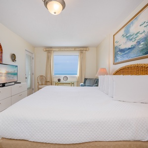 The master bedroom features a large bed, a TV, and a stunning view of the beach through the window