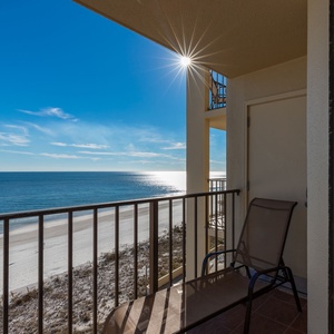 The balcony offers a serene view of the tranquil water and white sand beach, complemented by a sunlit sky