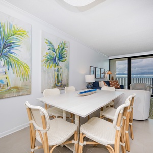 The bright dining area features a stylish white table, comfortable seating, and a stunning beach view through large windows