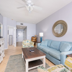 Bright living area featuring a blue sofa with queen sleeper, beach-themed chairs, and a balcony with a beach view