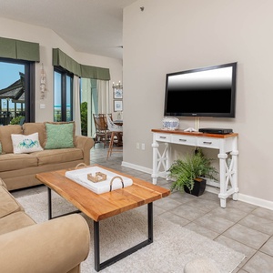 Living area features a plush sofa with decorative pillows, a wooden coffee table, and large windows offering a beach view