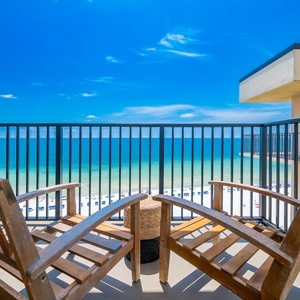 Relax on the balcony in wooden chairs, overlooking the serene beach and vibrant blue waters under a clear sky