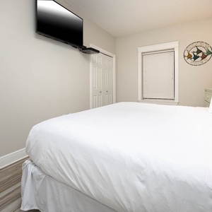 A comfortable guest bedroom features a large king bed with crisp white linens, a mounted TV, and a charming coastal decor