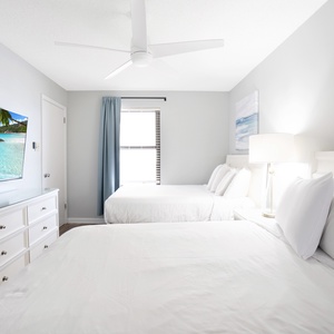 A bright guest bedroom features two double beds with crisp white linens, a large TV, and coastal artwork