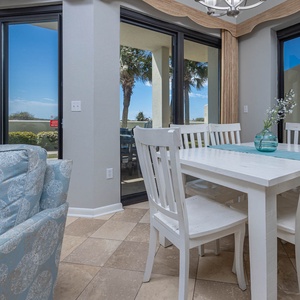 Bright dining area features a white table surrounded by chairs, with large windows offering a view of palm trees outside