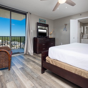 Bright master bedroom featuring a king bed, a wicker chair, a TV, and the balcony door leading to a scenic outdoor view