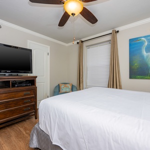 Cozy guest bedroom features a queen bed, a wooden dresser with a TV, and a colorful bird painting above the bed