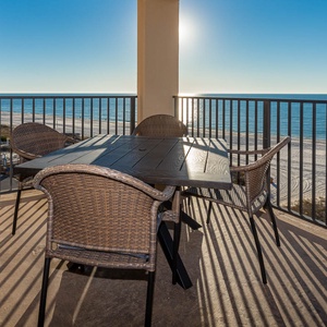 The balcony with a wooden table and wicker chairs offers a serene view of the beach and sparkling blue waters