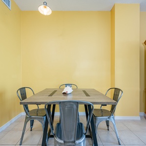 This dining area features a vibrant yellow wall, a wooden table, metal chairs, and a TV, with a view of the beach beyond