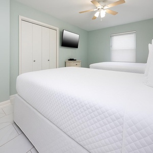 Bright guest bedroom featuring two queen beds, a wall-mounted TV, and light green walls, creating a refreshing retreat