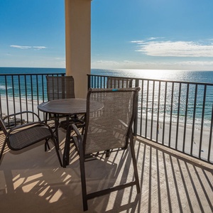 The balcony features a round table with four chairs, offering a serene view of the beach and sparkling water beyond