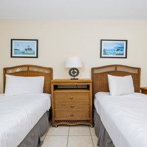 The bedroom features two twin beds with wicker headboards, complemented by coastal artwork and a charming nightstand
