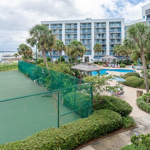 This view showcases a vibrant tennis court alongside a sparkling pool area with lush greenery and palm trees by the lagoon