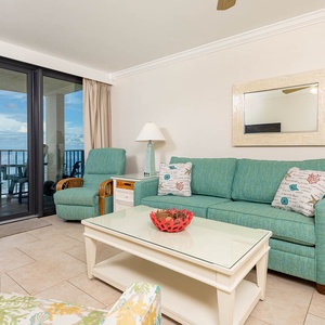 This living area features bright turquoise seating, a glass-top coffee table, and a breathtaking view of the beach