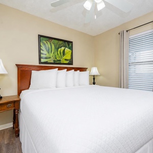 This bedroom features a large bed with crisp white linens, a tropical wall art piece, and wooden accents for a relaxing stay