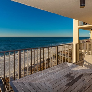 A spacious balcony features a wooden table and chairs, offering a serene view of the beach front and tranquil blue waters