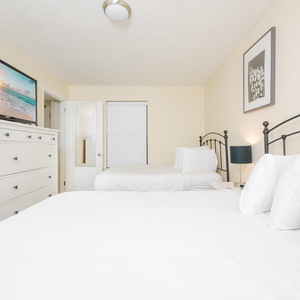 A cozy guest bedroom features two double beds, a large TV on a white dresser, and a soft color palette with minimal decor