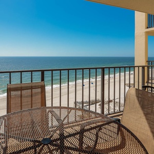 The balcony features a round table and chairs, offering a panoramic view of the beach and sparkling water below