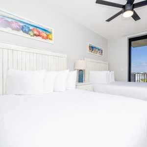 Relax in the airy guest bedroom featuring two comfortable double beds, coastal artwork, and a view from the balcony