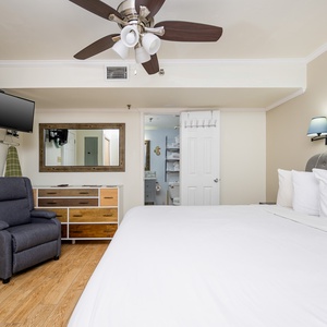 The room features a large king bed with crisp white linens, a cozy recliner, a wall-mounted TV, and a stylish dresser