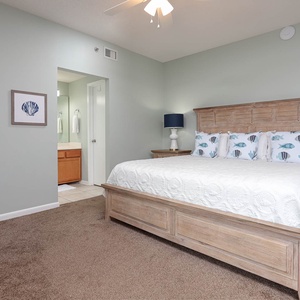 Master bedroom featuring a king bed with coastal-themed pillows and a soft carpet, leading to an adjoining bathroom
