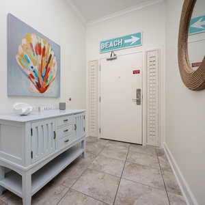 This welcoming entryway features a colorful shell painting, a light blue console table, and a sign pointing to the beach