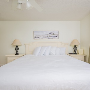 The master suite bedroom featuring a large king bed, bedside lamps, and a view of the beach through sliding glass doors
