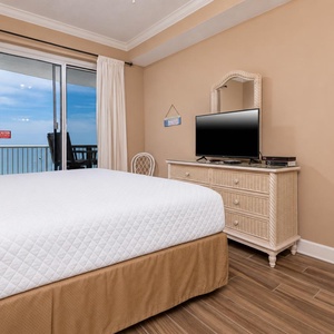 The bedroom features a comfortable bed, a TV for entertainment, and a stunning beachfront view from the sliding glass door