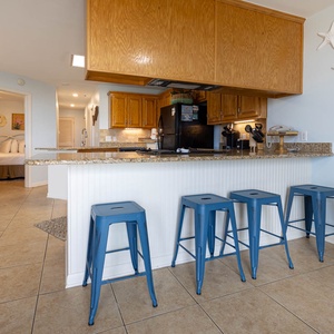 A cozy breakfast bar with blue stools complements the kitchen's wooden cabinetry, leading to a welcoming bedroom