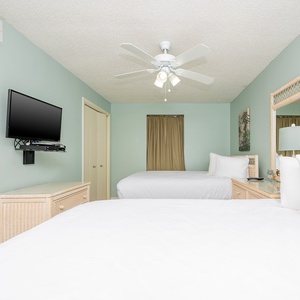 This guest bedroom features two neatly made queen beds, a wall-mounted TV, and soft green walls that create a calming vibe