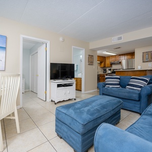 The living area features comfortable blue sofas, a dining table, and a view of the beach through the artwork