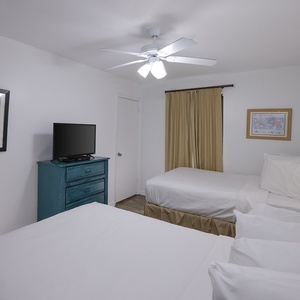 A bright guest bedroom features two double beds, a teal dresser with a TV, and soft lighting from a bedside lamp