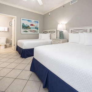 Brightly decorated guest room features two double beds with navy skirts, a bedside lamp, and a vibrant beach art on the wall