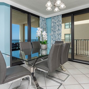 A stylish dining area features a glass table surrounded by gray chairs, with large windows showcasing a beach front view