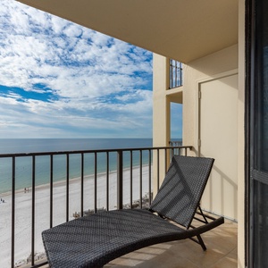 A black lounge chair sits on the balcony, offering a view of the calm waters and white sand below under a partly blue sky